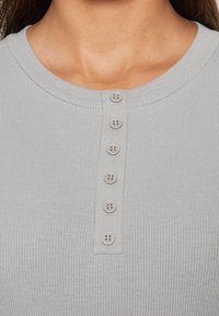 Top grigio chiaro a coste con scollatura tonda e sei bottoni abbinati sulla parte anteriore. Il tessuto sembra morbido con una texture liscia.