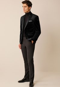 Black velvet blazer with a single pocket, worn over a black turtleneck and paired with slim-fit black trousers and glossy boots.
