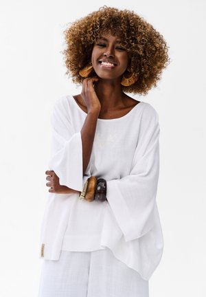 Uśmiechnięta kobieta o kręconych włosach nosi duże kolczyki, drewniane bransoletki oraz luźną białą bluzkę z długim rękawem i pasujące spodnie na jednolitym tle.