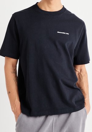Man wearing plain black short-sleeve t-shirt with small white "Abercrombie | NYC" logo on left chest, hands in gray pants pockets.