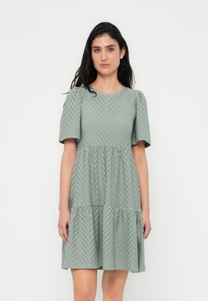 Groene jurk met korte mouwen, een chevronpatroon en een gelaagde rok. Gemaakt van een getextureerde stof met uitsnijdingen. Getailleerd lijfje.