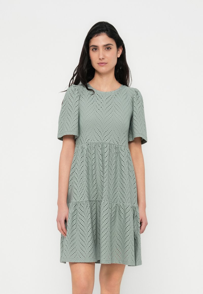 Zielona sukienka z krótkim rękawem, z wzorem w kształcie jodełki i spódnicą z falbanami. Wykonana z fakturowanej tkaniny z wycięciami. Dopasowany gorset.