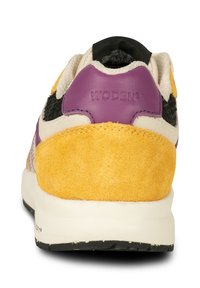 Sneaker vu de dos avec talon en daim jaune, col en cuir violet, tissu noir et beige, et semelle blanche avec semelle extérieure noire.