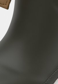 Gros plan sur une botte de pluie en caoutchouc gris foncé avec une semelle texturée sur une surface blanche et une étiquette en carton brun attachée.