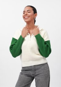 Light-colored, ribbed sweater with a green oversized, textured sleeve and half-zip front. Worn with gray jeans, featuring a relaxed fit.