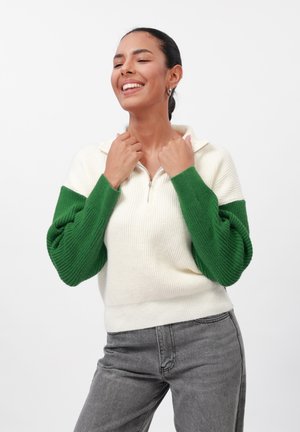 Pull léger à côtes de couleur claire avec une manche verte oversized et texturée, et un zip demi-long. Porté avec un jean gris, d'ajustement décontracté.
