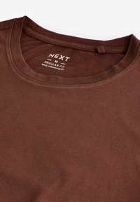 Brun bomulds T-shirt med rund hals, der har et label med hvid tekst, der angiver mærke og størrelse, med en glat overflade.