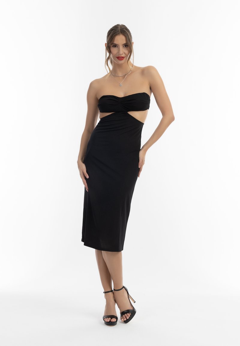 Schwarzes, trägerloses Midikleid mit einem geknoteten Bustier-Design und ausgeschnittenen Seiten, hergestellt aus einem glatten, elastischen Stoff. Kombiniert mit schwarzen High Heels.