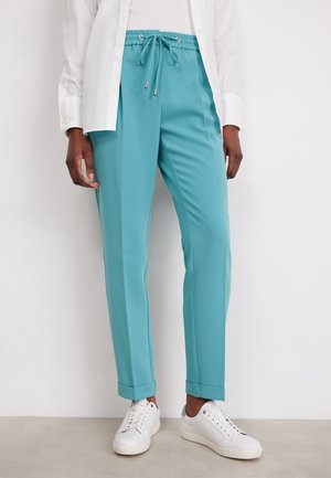 Turkusowe spodnie ze ściągaczem i podwiniętymi nogawkami, noszone z białymi trampkami i białą koszulą zapinaną na guziki.