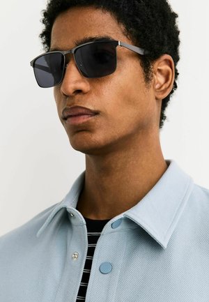 Ung mand iført sorte rektangulære solbriller og en lyseblå jakke med krave over en sort-hvid stribet skjorte.