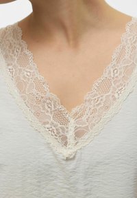 Top bianco con scollatura a V profonda e delicata bordatura in pizzo avorio, che mette in mostra motivi floreali lungo i bordi e un tessuto morbido e strutturato.