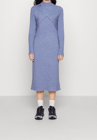 Vestido de punto azul con canalé, de manga larga y cuello alto, que presenta un diseño cruzado angular en el pecho, combinado con zapatillas negras.