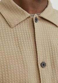 Šviesiai ruda tekstūruota marškiniai su tinklelio raštu, turintys smailų apykaklę ir vieną juodą sagą. Minkšta medžiaga su audinio išvaizda.