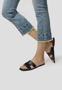 Zwarte leren slidesandalen met studs en een open teen ontwerp, gecombineerd met lichtblauwe geborduurde jeans met een franjes zoom.