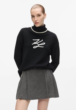 Kvinde iført en sort højhalset sweater med hvide kanter og initialer, kombineret med et plisseret gråt skørt, stående på en hvid baggrund.