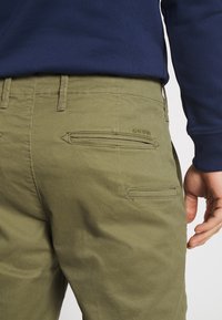 Olivgröna byxor med texturerad tyg, med två bakfickor med dragkedja och en logotyp på sidan. Bärs med en marinblå topp.