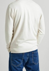 Mann trägt ein cremefarbenes Langarmshirt und blaue Jeans, von hinten vor einem einfarbigen hellen Hintergrund gezeigt.