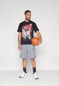 Czarny graficzny t-shirt z czerwono-białym napisem "BULLS", szare spodenki sportowe, białe tenisówki i trzymający pomarańczową piłkę do koszykówki.