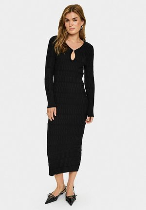 Black ribbed knit dress with long sleeves, v-neckline featuring a keyhole detail, and body-hugging silhouette, paired with pointed-toe shoes.