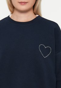 Felpa blu navy realizzata in tessuto morbido, con un design a forma di cuore in strass sulla parte sinistra del petto. Collo tondo e vestibilità rilassata.