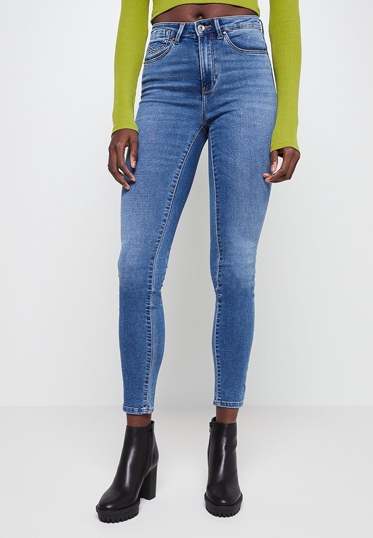 Jean skinny bleu associé à des bottines noires à talons et un haut court à manches longues vert citron sur une personne debout.