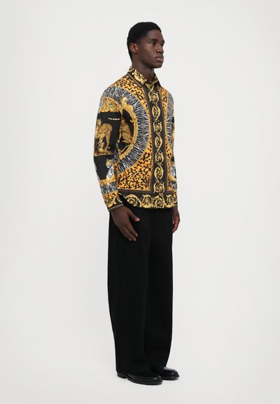 Cămașă cu model negru și auriu, cu motive animale, detalii complexe, mâneci lungi și guler, purtată cu pantaloni negri.