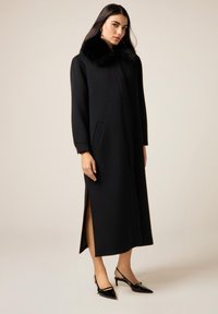 Cappotto nero in lana con colletto in pelliccia sintetica, maniche lunghe, spacco laterale e tasche laterali; abbinato a décolleté nere con punta appuntita.