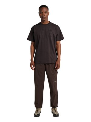 Jeune homme debout, portant un t-shirt noir à manches courtes, un pantalon marron foncé avec une poche zippée et des chaussures de randonnée beiges avec des semelles noires.