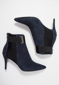 Bottes cheville en daim bleu marine avec un bout pointu, des panneaux latéraux élastiques noirs et un détail métallique sur la sangle de cheville.
