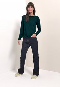 Grünes, gemustertes Langarmshirt, dunkelblaue ausgestellte Jeans und hellfarbene Turnschuhe. Der Stoff wirkt glatt und das Design ist leger geschnitten.