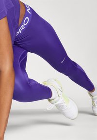Fioletowe legginsy z białym logo "Nike" oraz pasem w kolorze "PRO", w połączeniu z białym i jasnoniebieskim obuwiem sportowym. Gładki, elastyczny materiał.
