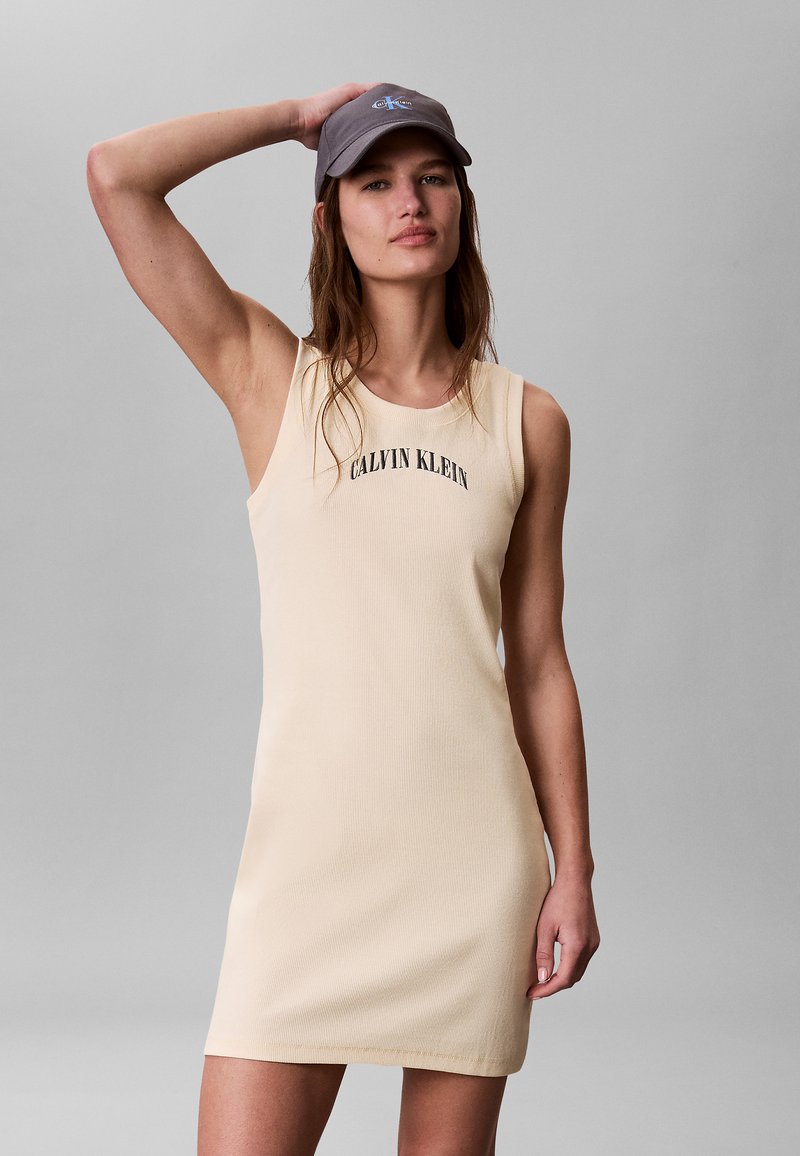 Femme portant une robe beige sans manches Calvin Klein et une casquette grise, posant avec une main touchant l'arrière de sa tête.