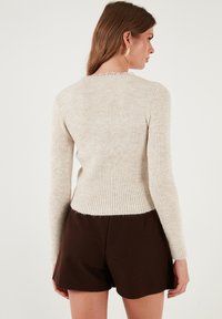 Bežinė raukinėta megztinė su ilgomis rankovėmis, turinti prigludusią juosmenį ir tekstūruotą audinį. Šviesesnis atspalvis apykaklėje, dėvima su rudomis šortais.