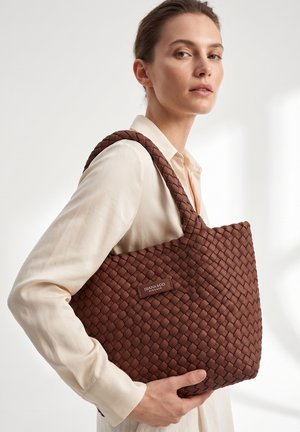 Vrouw met een crème blouse houdt een grote bruine gevlochten leren shopper met dikke handvatten en een merkpatch op de schouder vast.