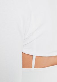 Bílý krátký top z měkké látky, s bočním výstřihem a tenkým páskem pod ním. Hladká textura, neformální design.