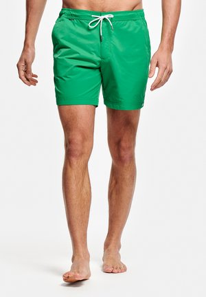 Man draagt felgroene zwembroek met witte trekkoord, loopt op blote voeten tegen een witte achtergrond, torso en benen zichtbaar.