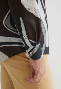 Chemise à manches longues avec une base marron et des motifs abstraits blancs et noirs, dotée de poignets à boutons et d'une texture de tissu lisse.