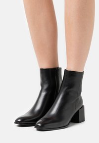 Black leather ankle boots with a square toe, mid-height block heel, and side zipper detail. Smooth texture with minimalist design.