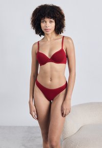 Κόκκινο εσώρουχο σετ που περιλαμβάνει ένα λείο, μαξιλαρισμένο σουτιέν με ρυθμιζόμενους ιμάντες και ταιριαστά ψηλόμεσα σλιπ. Το ύφασμα φαίνεται μαλακό και ελαστικό.