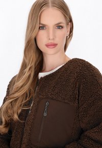 Brown faux fur jacket with a textured body, a zippered chest pocket, and a soft, cozy design. Features zippered closure and contrast panels.