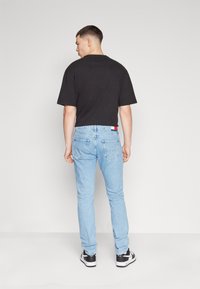 Camiseta negra de manga corta y jeans de mezclilla azul claro con un corte ajustado. Los jeans tienen una etiqueta roja y blanca en la cintura trasera. Zapatillas negras.