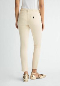 Pantaloni slim-fit de culoare crem, cu un brâu împletit și două buzunare la spate, asortați cu pantofi plata metalici aurii.