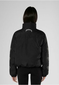 Sort pufferjakke med høj krave, elastiske manchetter og hvide broderede detaljer på ærmerne. Glat tekstur, cropped design.