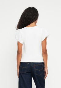 Femme aux longs cheveux bouclés portant un haut blanc à manches courtes et un jean bleu foncé, se tenant de dos devant un fond uni.