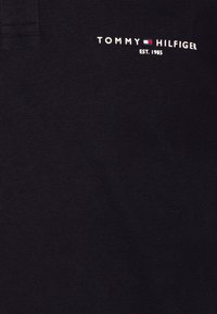 Černá bavlněná látka s texturovaným povrchem, na které se nachází bílý logo "TOMMY HILFIGER" a detail "EST. 1985" v kontrastu.