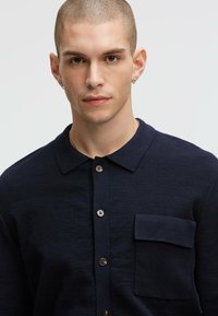 Marinefarbener Strickshirt mit Kragen, Knopfleiste und einer einzelnen Brusttasche. Verfügt über braune Knöpfe und einen strukturierten Stoff.