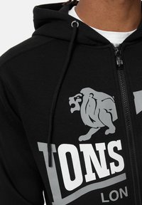 Zwarte hoodie met rits, gemaakt van zacht materiaal, met een opvallende grijze leeuwafbeelding en vetgedrukte witte tekst. Inclusief trekkoorden en een ritssluiting.