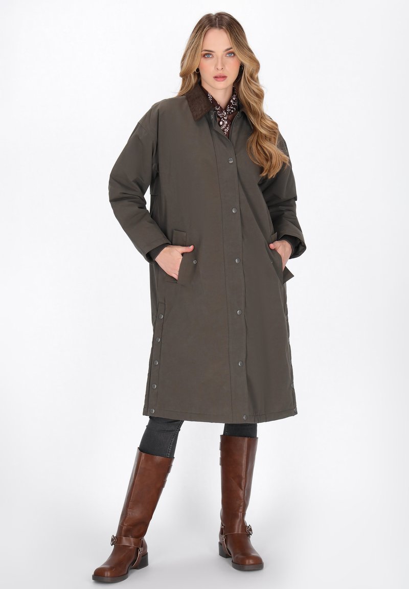 Cappotto lungo verde oliva con colletto marrone, bottoni a pressione, tasche laterali e silhouette dritta, abbinato a stivali marroni al ginocchio.
