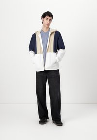 Lichte, kleurgeblokte jas in marineblauw, beige en wit; heeft een capuchon, volledige rits en een relaxte pasvorm. Gecombineerd met donkere wijde jeans.