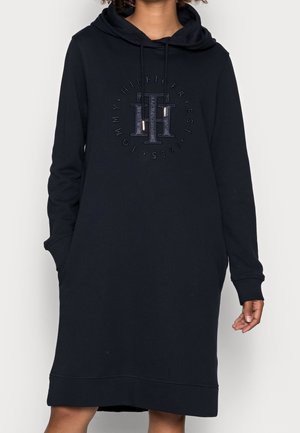 Femme portant une longue robe à capuche noire avec des poches avant et le logo Tommy Hilfiger brodé sur la poitrine.